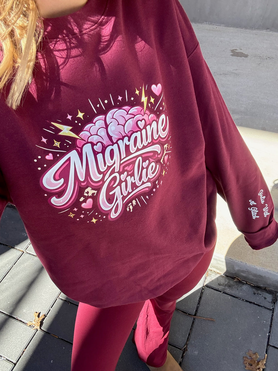 Maroon sweatshirt with 'Migraine Girlie' design on the front and Girlie with a Glitch on the sleeve, worn by a person on a sidewalk.