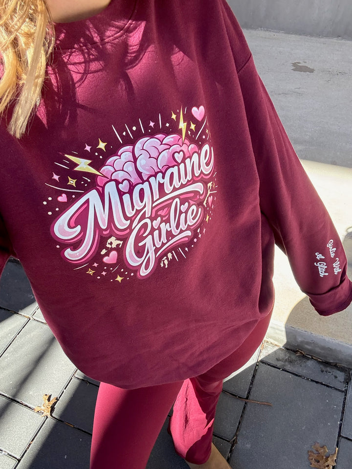 Maroon sweatshirt with 'Migraine Girlie' design on the front and Girlie with a Glitch on the sleeve, worn by a person on a sidewalk.