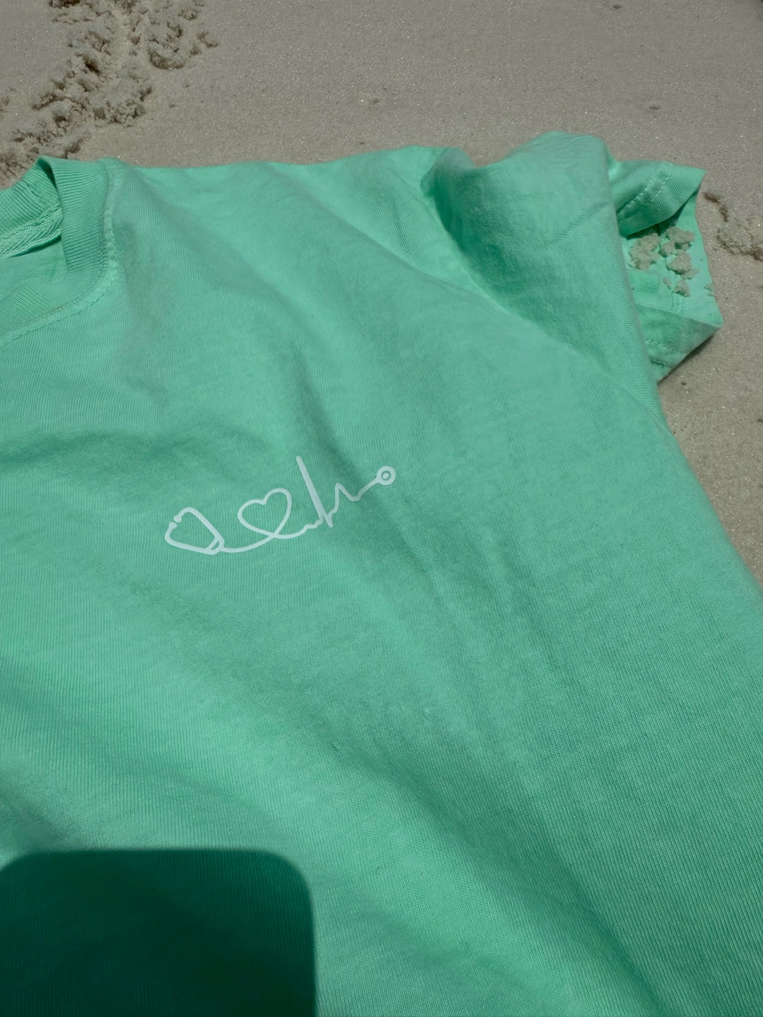 Close up of light green Heart of Medicine t-shirt featuring stethoscope and heart waveform logo on the upper left chest, displayed on sandy beach.