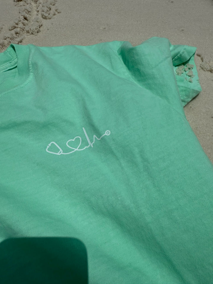 Close up of light green Heart of Medicine t-shirt featuring stethoscope and heart waveform logo on the upper left chest, displayed on sandy beach.