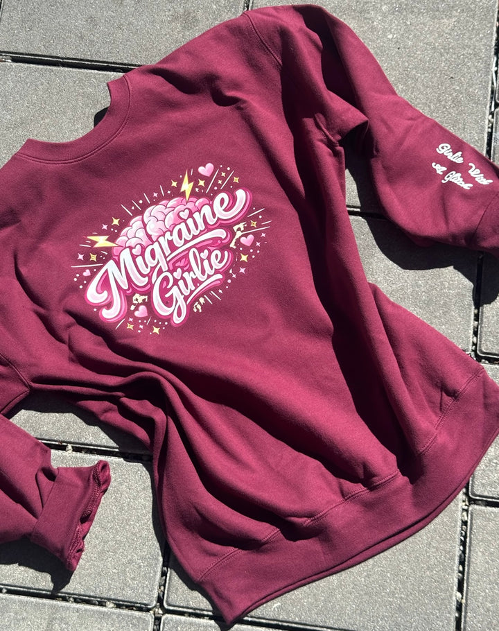 Maroon sweatshirt with 'Migraine Girlie' text and Girlie with a Glitch on the sleeve laying on a tiled floor