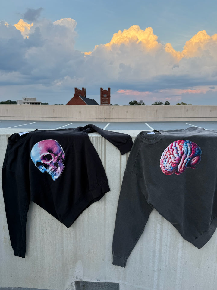 Two black sweatshirts on a concrete wall, one with a colorful skull, the other with a colorful brain