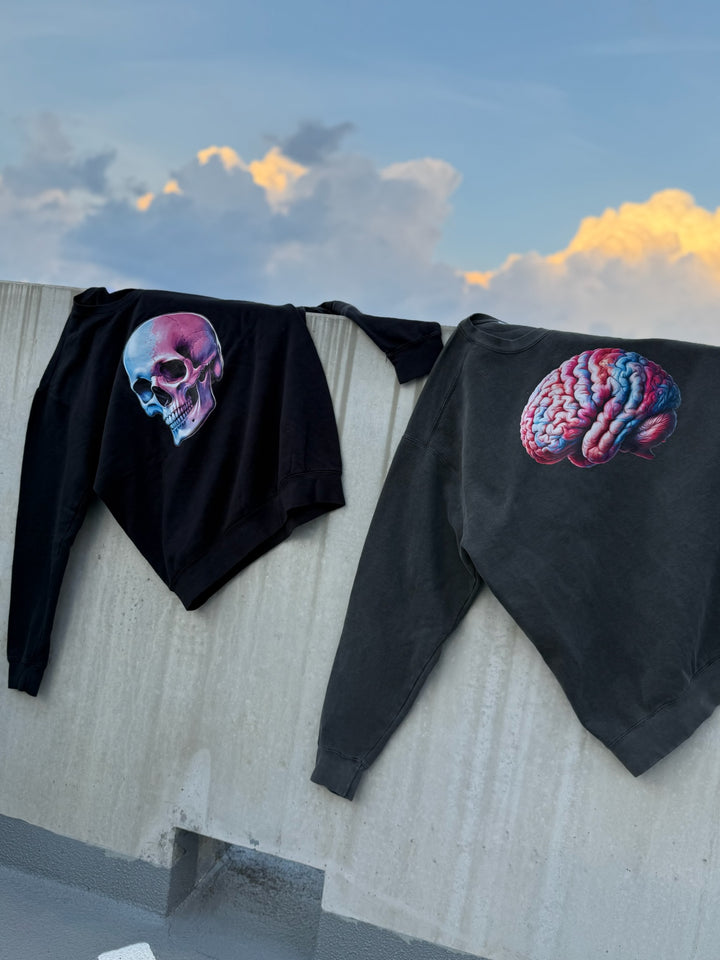 Two black sweatshirts on a concrete wall, one with a colorful skull, the other with a colorful brain