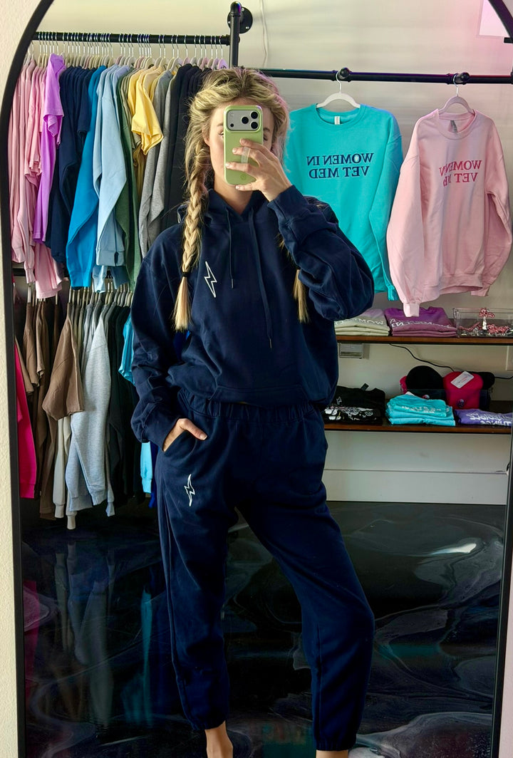 Person wearing a navy tracksuit taking a mirror selfie in a room with clothes on racks.