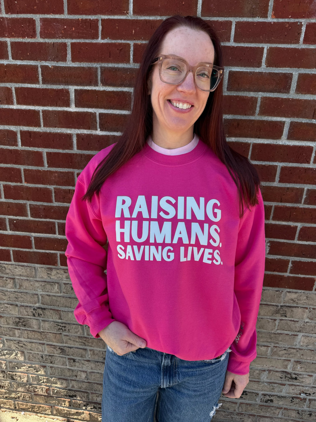 Ladyspinedoc wearing a hot pink crewneck sweatshirt with the text 'RAISING HUMANS. SAVING LIVES.' printed on the front.