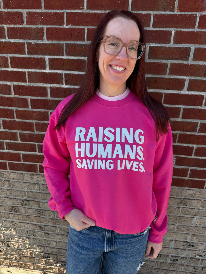 Ladyspinedoc wearing a hot pink crewneck sweatshirt with the text 'RAISING HUMANS. SAVING LIVES.' printed on the front.