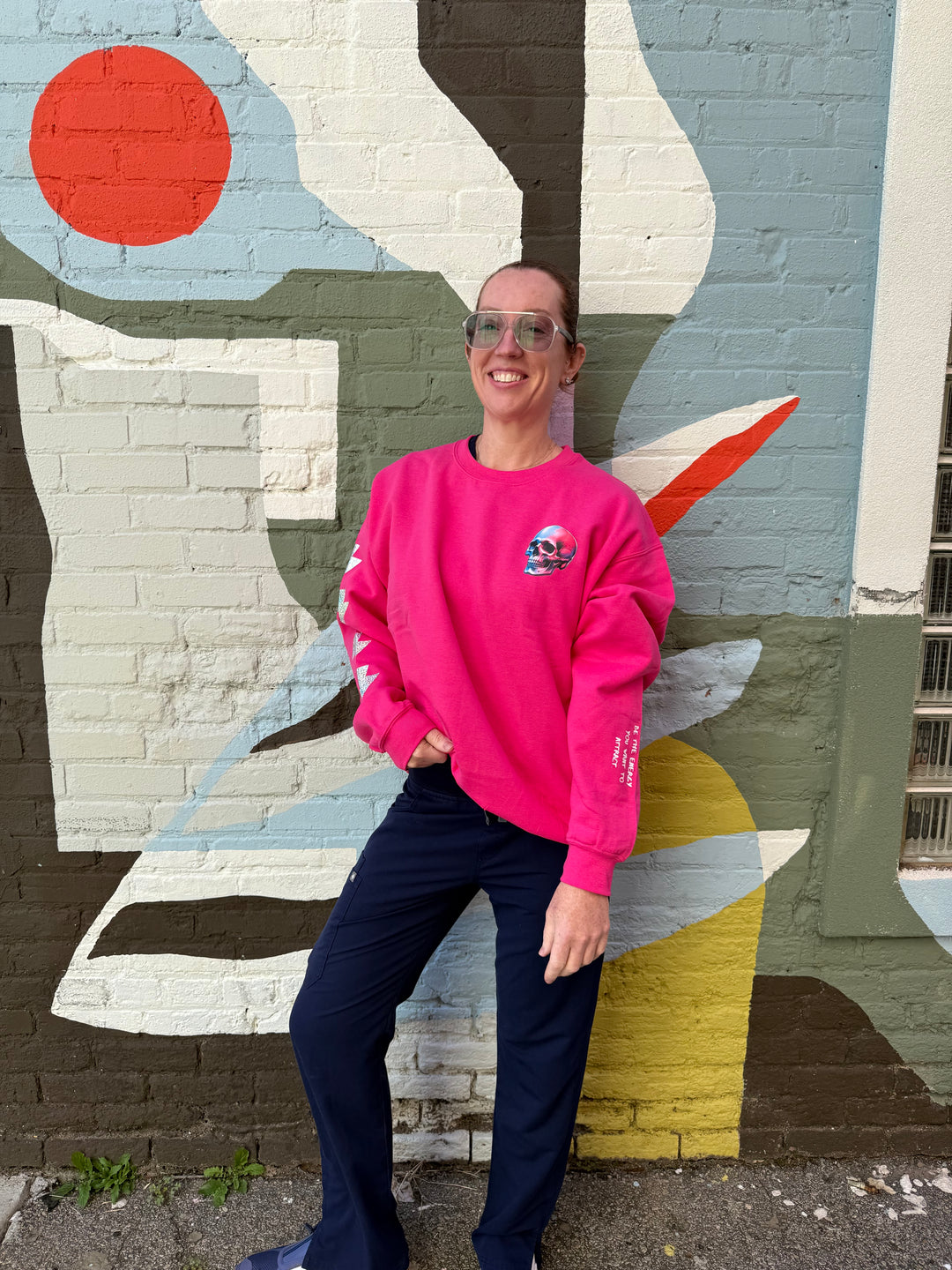 Ladyspinedoc wearing the The Power Within Crewneck Sweatshirt - Pink Shirt with Lightning Bolts, Skull, and sleeve art standing in front of a colorful abstract mural.