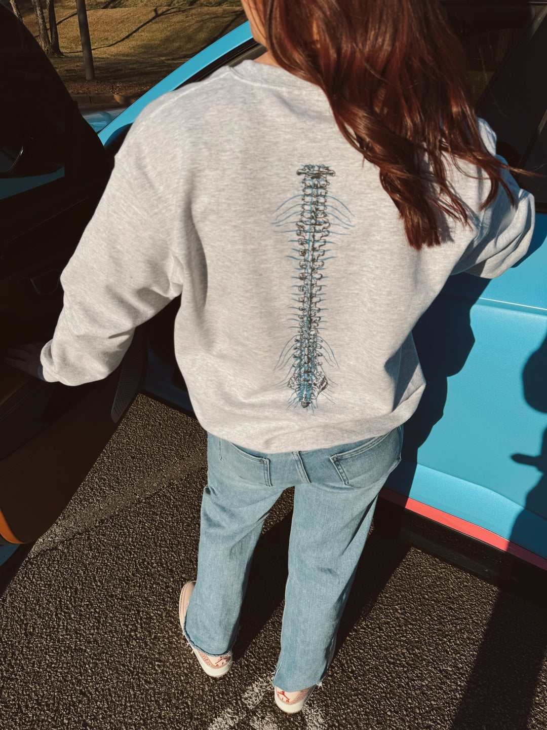 A person wearing a gray crewneck sweatshirt with a spine design on the back, standing next to a blue vehicle.