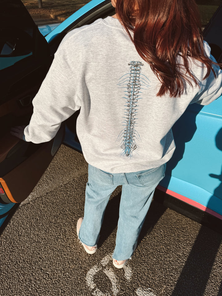 A person wearing a gray crewneck sweatshirt with a spine design on the back, standing next to a blue vehicle.