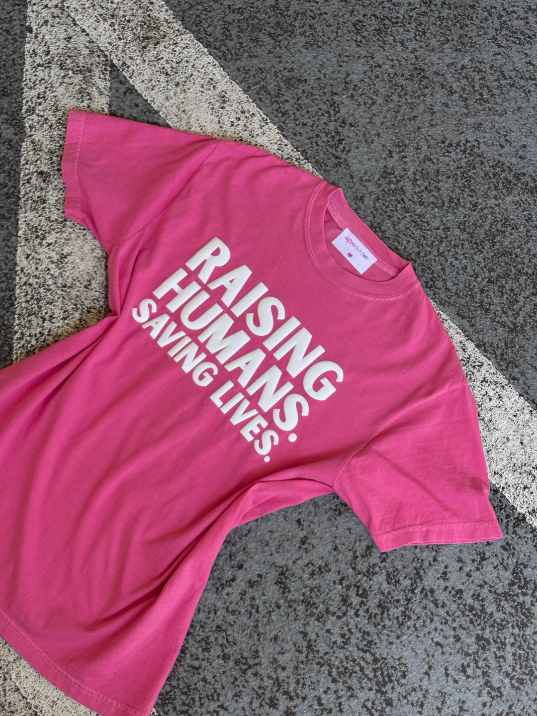 Pink t-shirt with text 'Raising Humans. Saving Lives.' on a textured surface.
