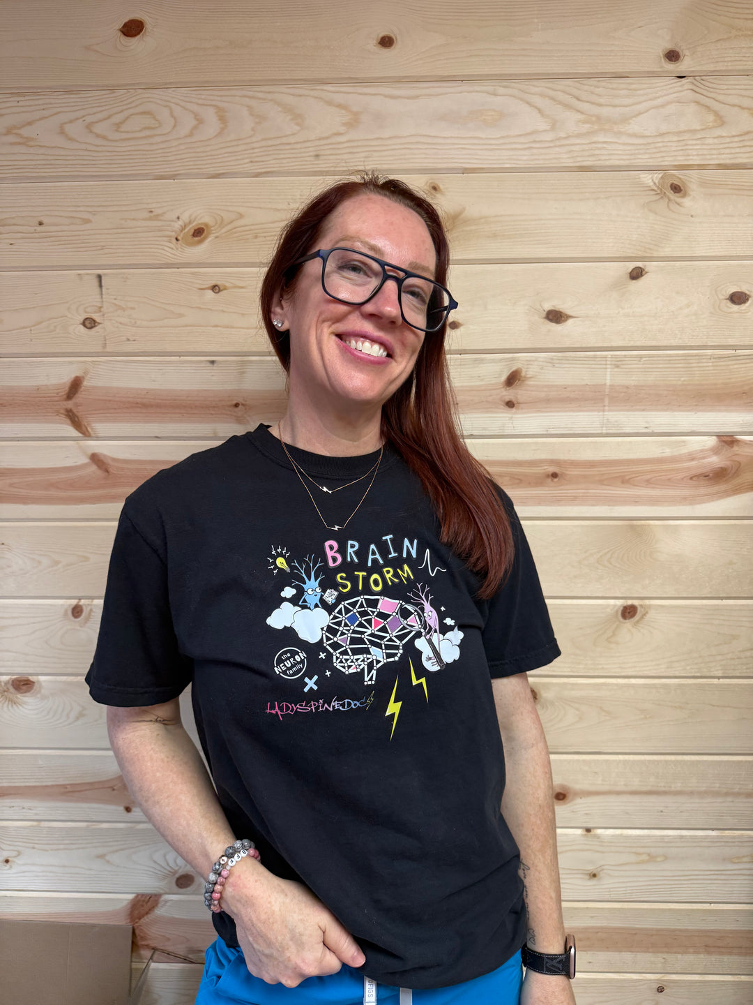 Ladyspinedoc wearing a black brain storm tee with colorful graphics against a wooden wall.