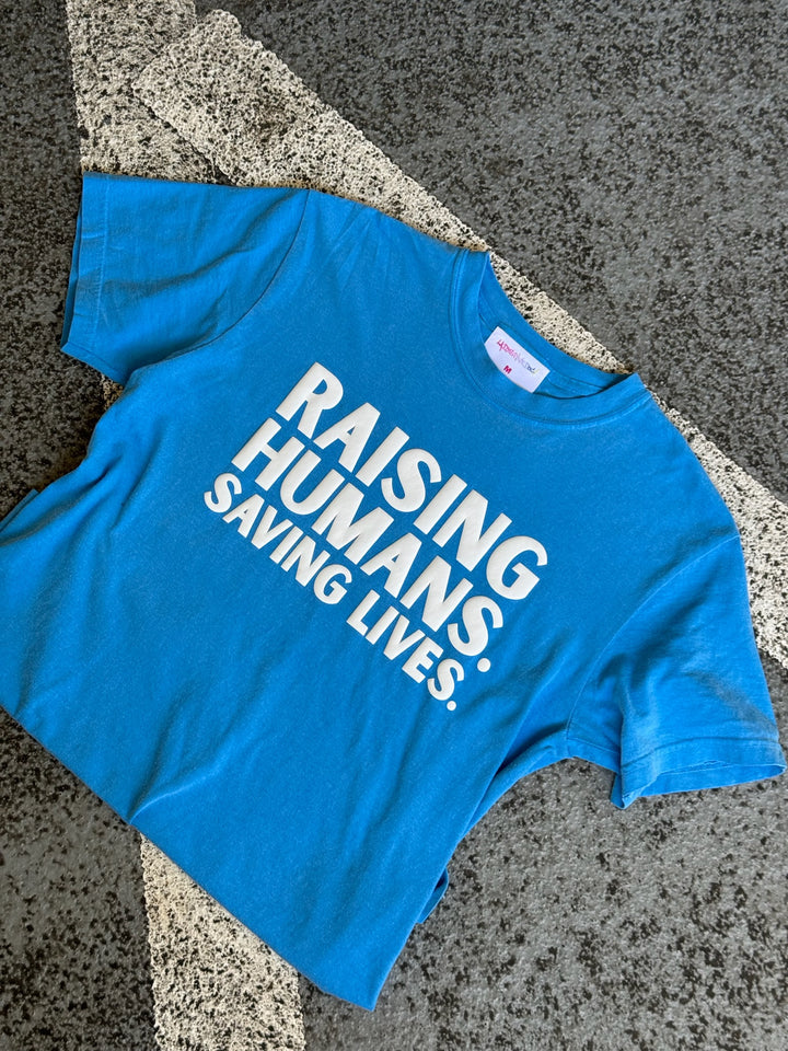 Blue t-shirt with text 'Raising Humans. Saving Lives.' on a textured surface.
