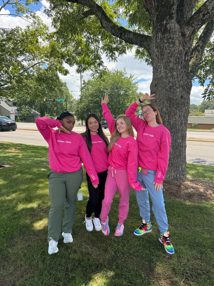 Four women in pink Caffeine + Botox sweatshirts posing outdoors near a tree