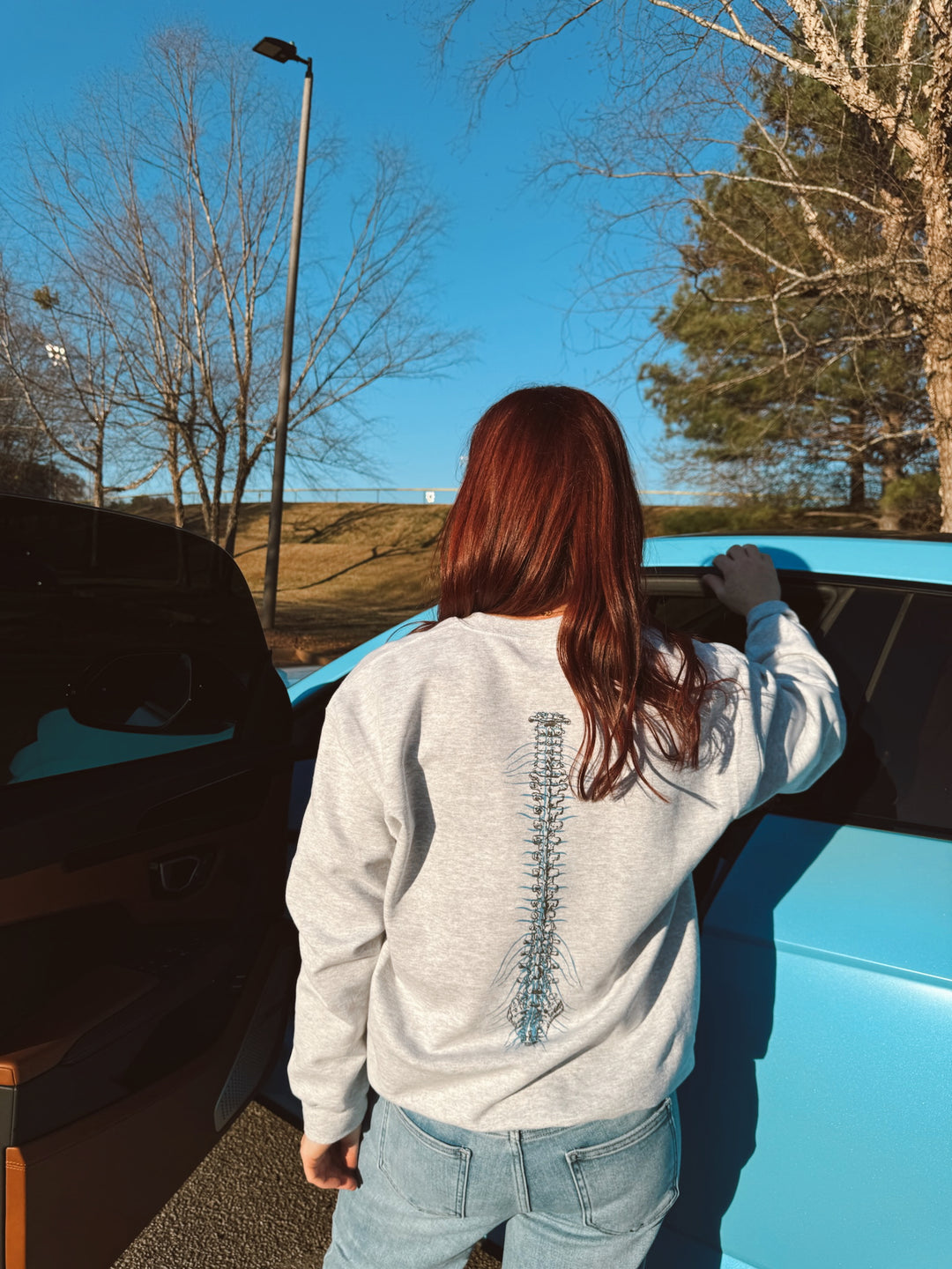 A person wearing a gray crewneck sweatshirt with a spine design on the back, standing next to a blue vehicle.