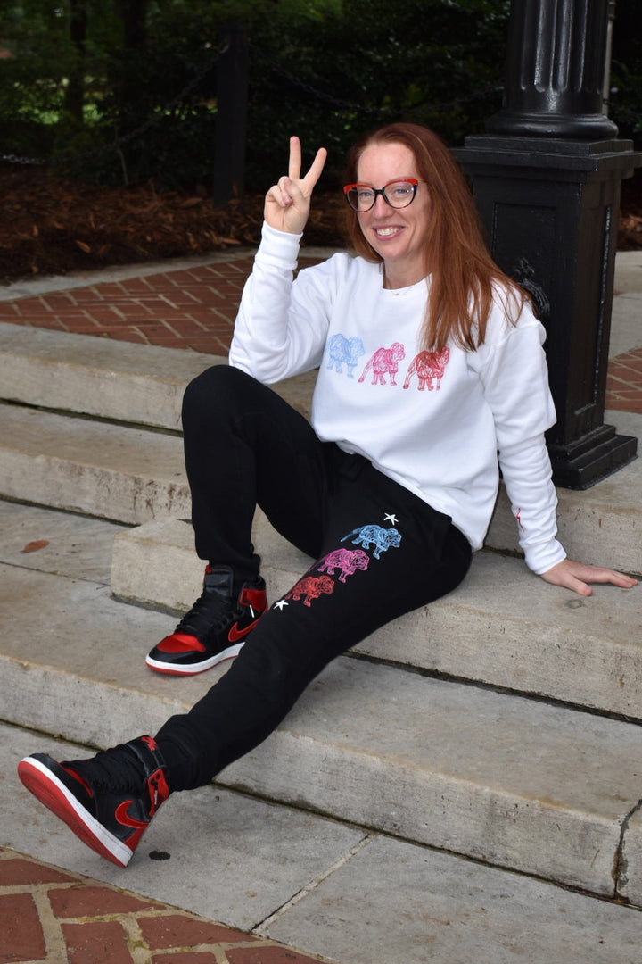Person sitting on steps wearing a white sweatshirt with colorful bulldog designs and black pants.