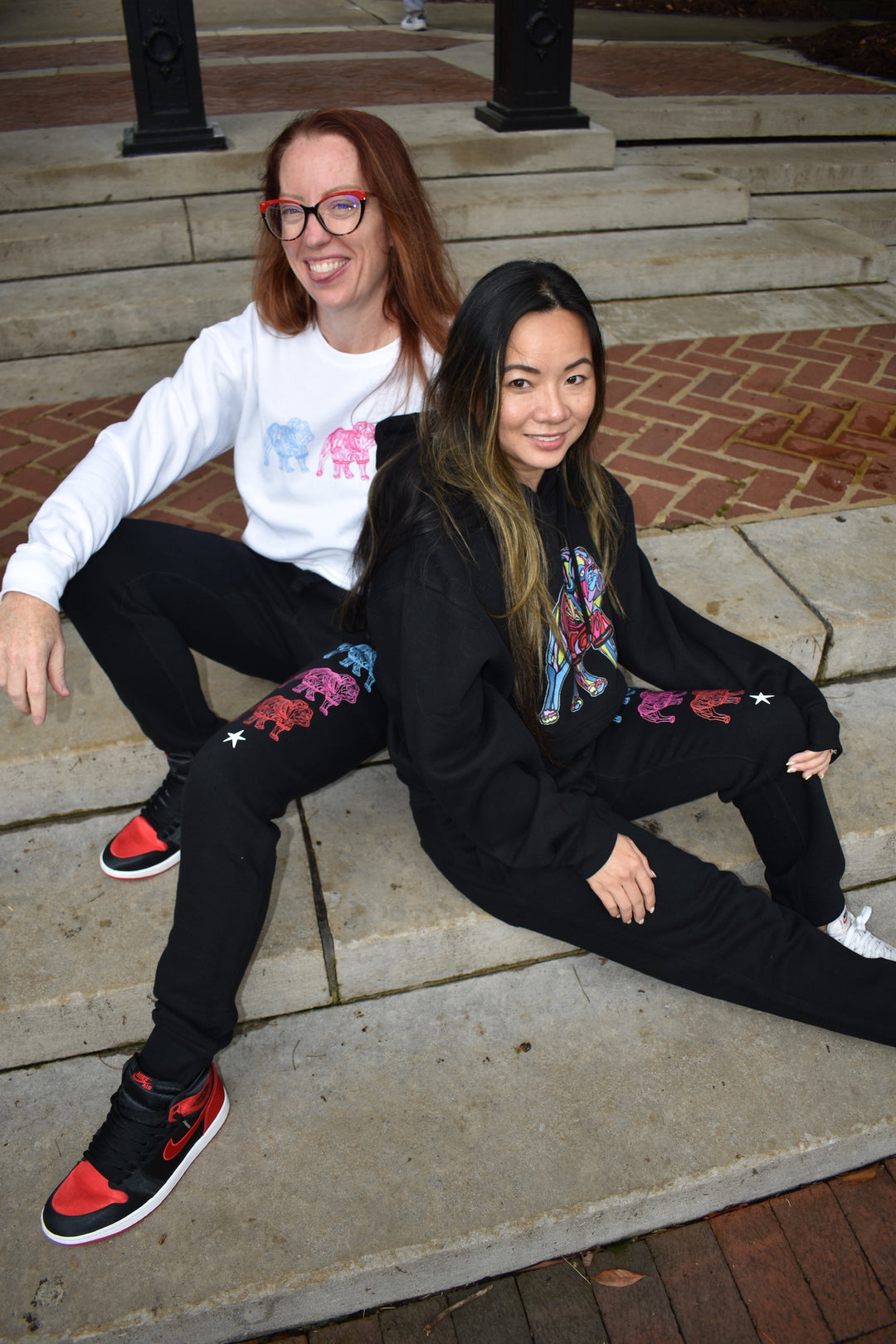 Two people sitting on steps outdoors, one wearing a white sweatshirt and black pants with colorful designs, the other in a black hoodie and pants.