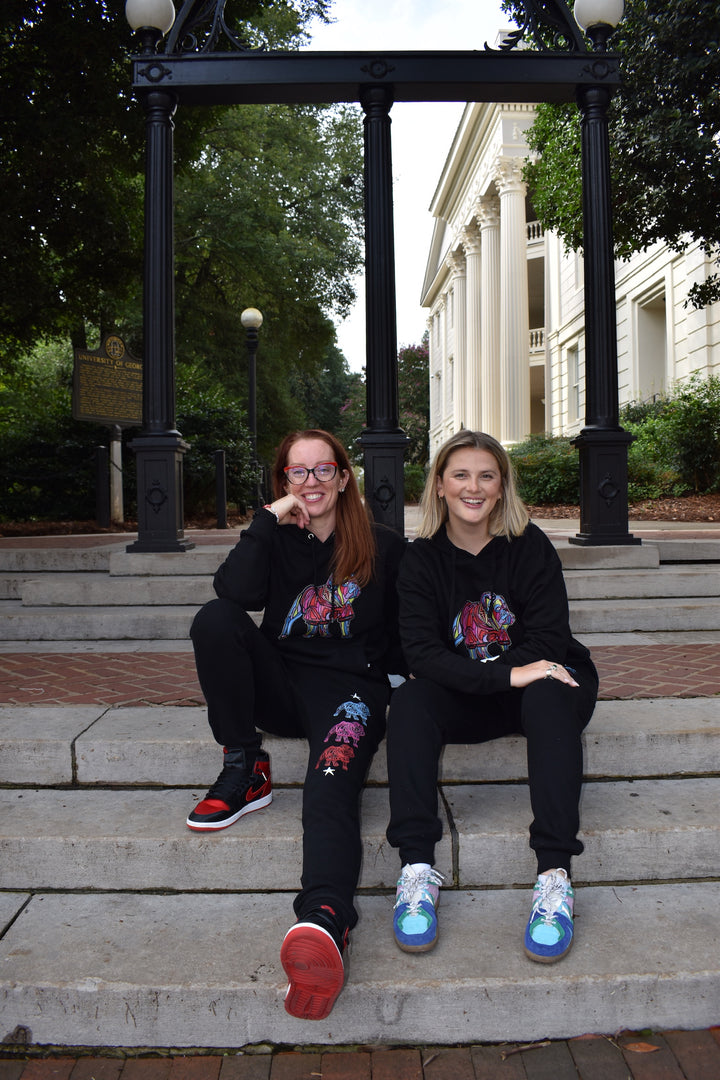 Two women wearing Chan Art Collab Game Day bulldog gear sitting on steps in front of a building with trees and a lamp post in the background.