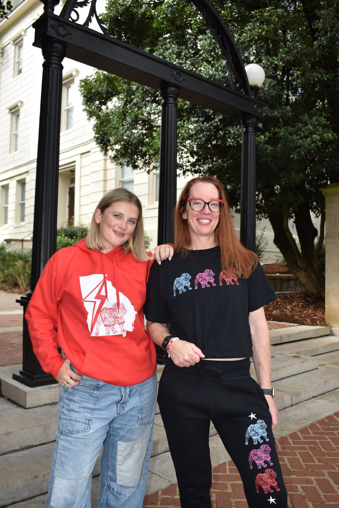 Two women wearing bulldog shirts standing together in front of a black metal archway with trees and a building in the background.
