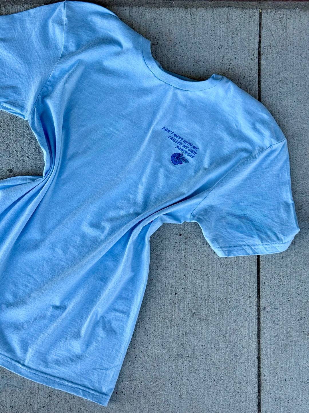 Front flat lay of a light blue t-shirt with "Don't Mess With Me, I Killed My Own Pancreas" logo on a concrete surface