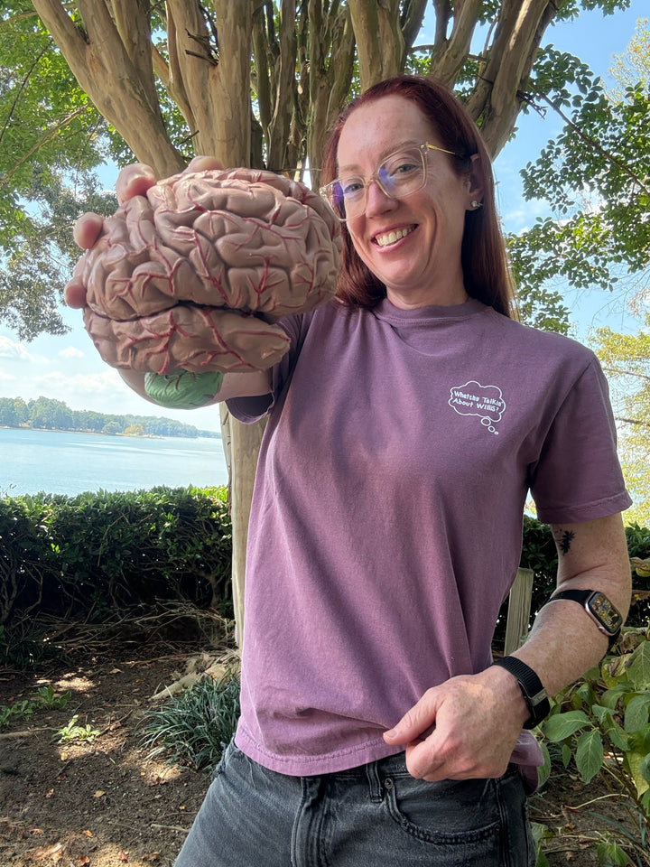Dr. Betsy Grunch wearing a Whatchu Talkin’ About Willis T-Shirt and holding a model of a brain outdoors near a body of water