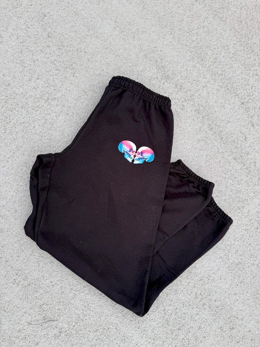 Super Skull Sweatpants - Folded Black Joggers with Skull Design laying on concrete