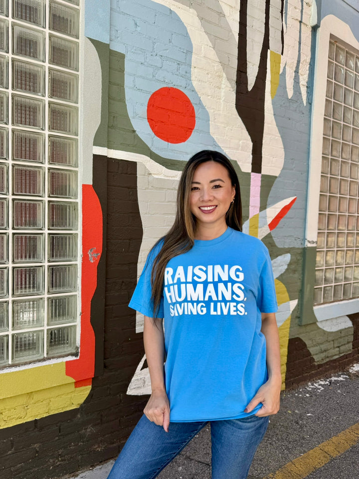 Woman wearing blue Raising Humans. Saving Lives. tee by Ladyspinedoc, standing in front of a mural with a bold, proud stance and smile.
