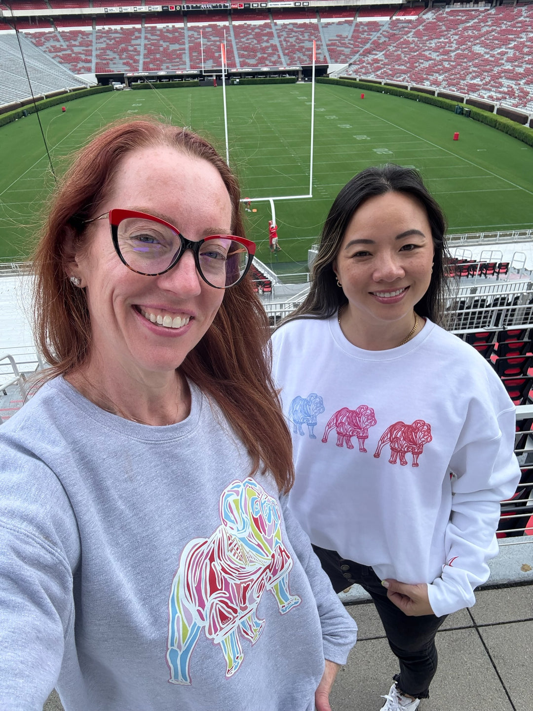 Two women wearing sweatshirts with colorful bulldog graphics in a stadium setting.