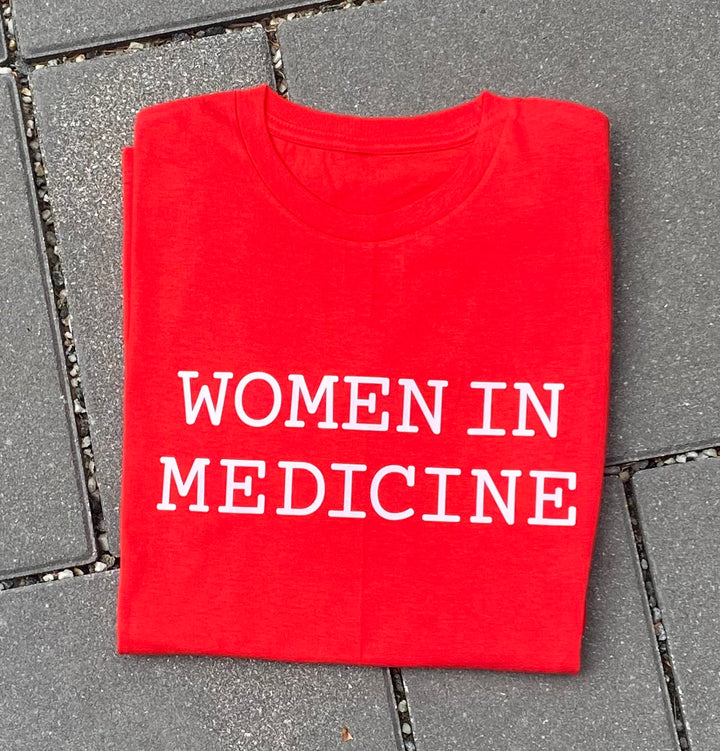 Folded red t-shirt with 'WOMEN IN MEDICINE' text on a gray pavement.