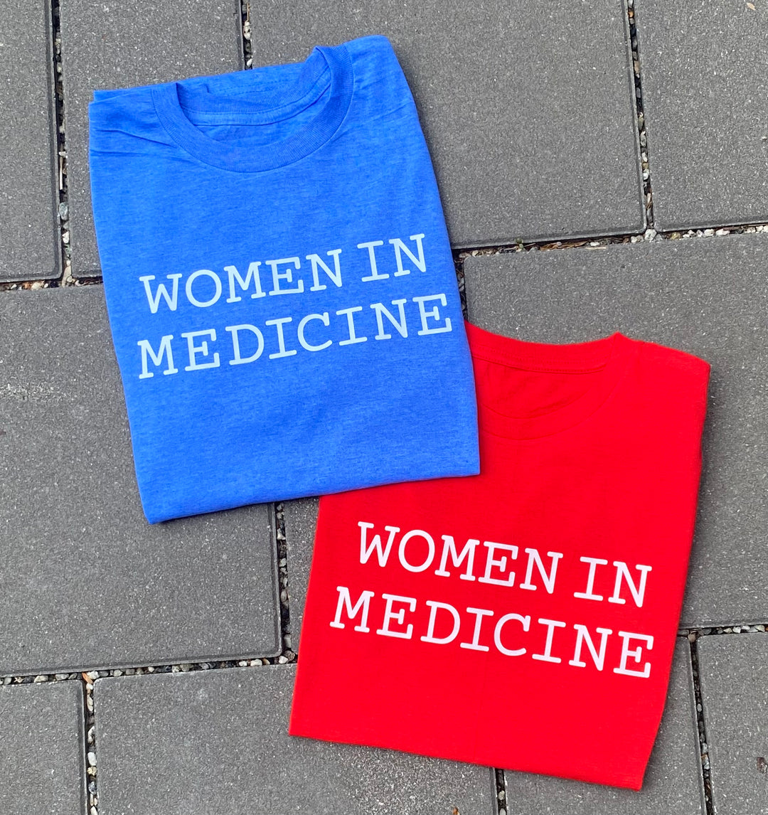 Blue and red t-shirts with 'WOMEN IN MEDICINE' text on a tiled floor.