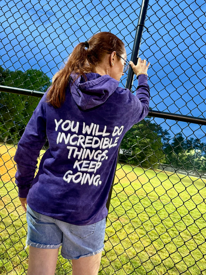 Dr. Betsy Grunch (Ladyspinedoc) wearing the purple Incredible Hoodie, displaying the back of the shirt showing bold white brushstroke message: “YOU WILL DO INCREDIBLE THINGS. KEEP GOING.”