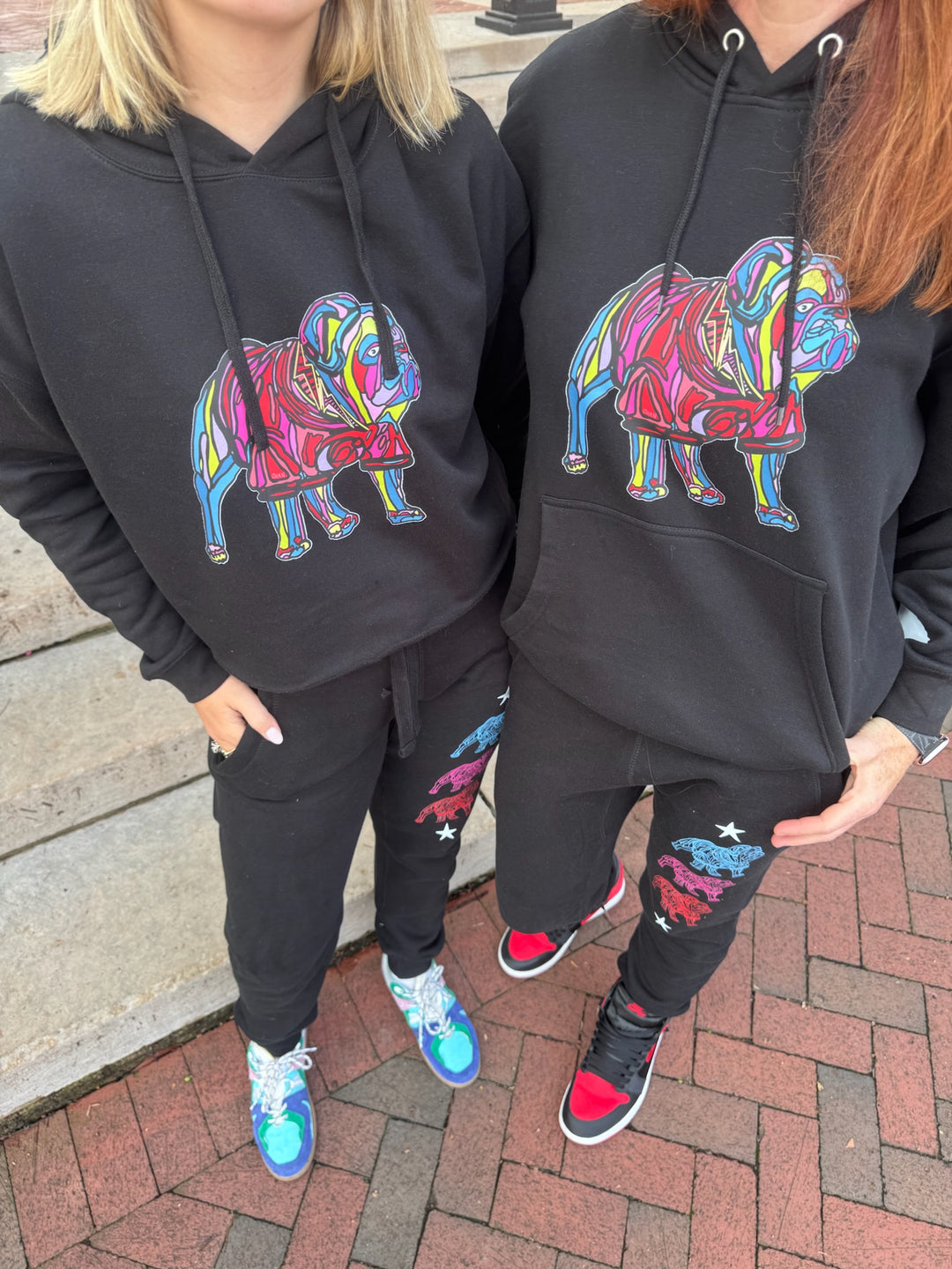 Two people wearing black hoodies with colorful bulldog designs on a brick pavement.