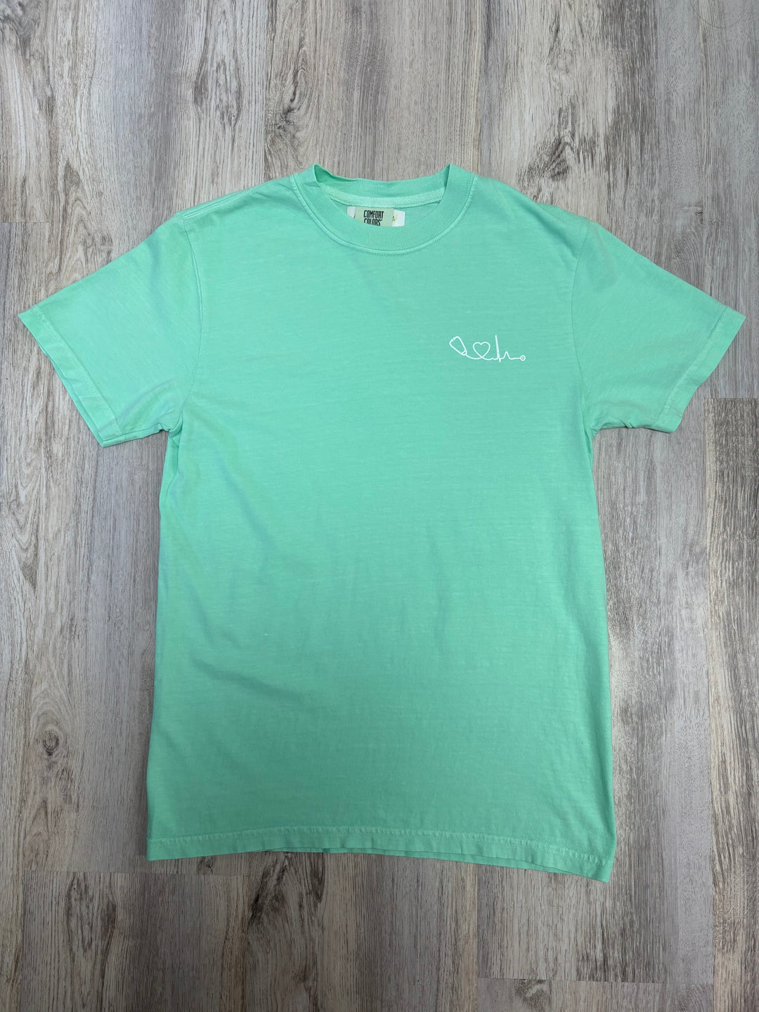 Flat lay of light green Heart of Medicine t-shirt featuring stethoscope and heart waveform logo on the upper left chest.