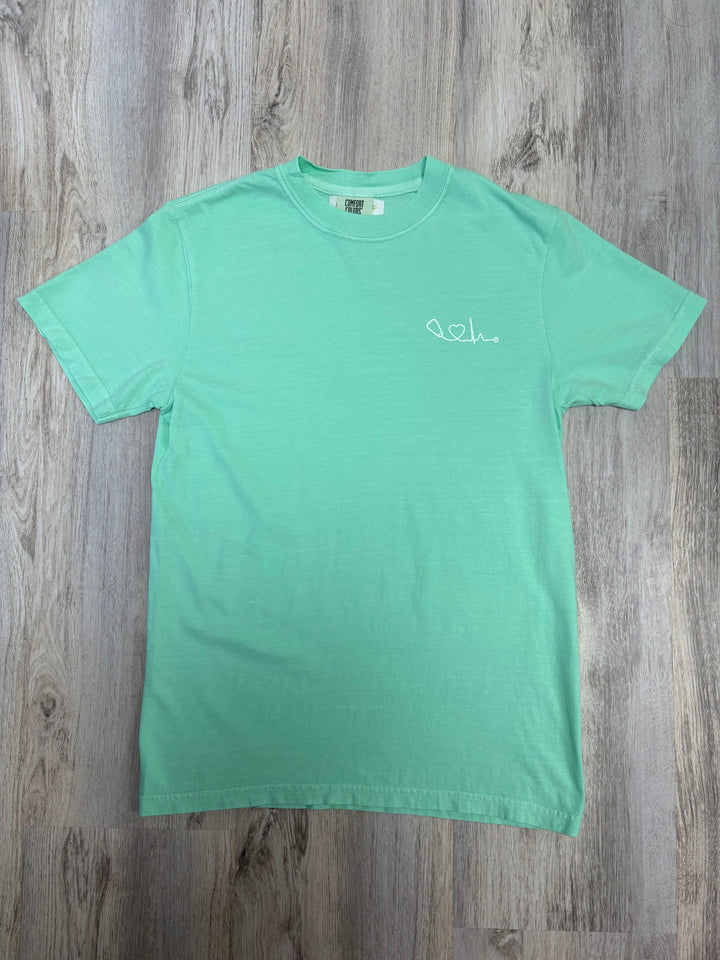 Flat lay of light green Heart of Medicine t-shirt featuring stethoscope and heart waveform logo on the upper left chest.