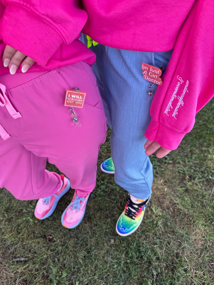 Two people wearing I Will Cut You and I'm Just A Girl Doctor badge reels with pink and blue clothes and colorful shoes on grass