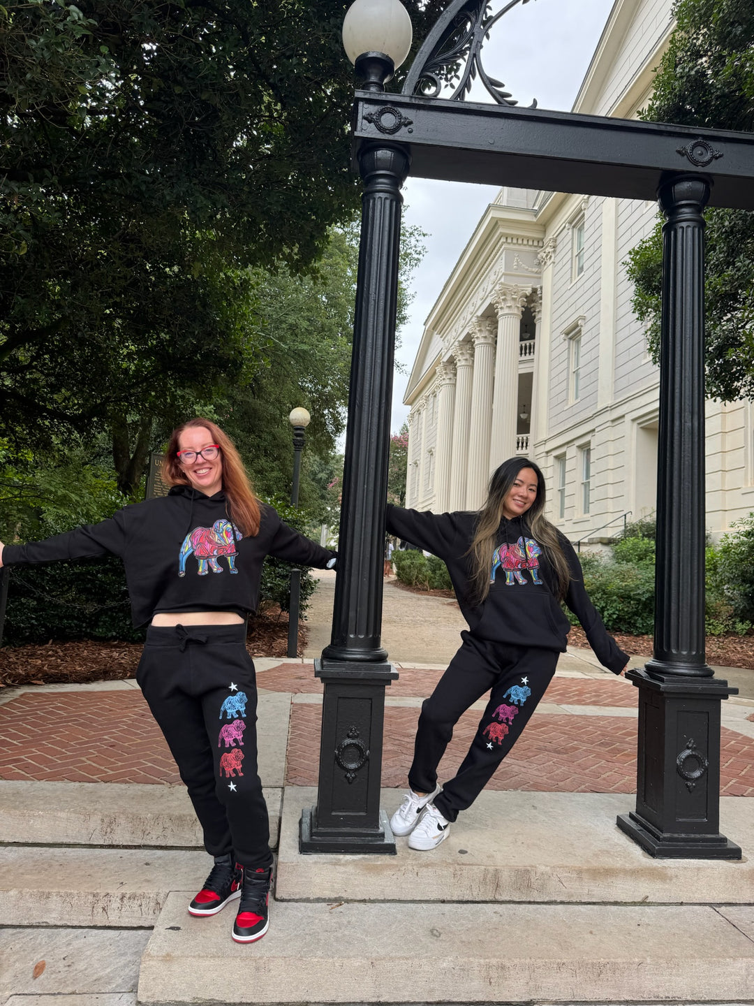 Two people wearing bulldog game day gear and posing outdoors with a building and trees in the background